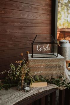 a wooden table topped with a glass box filled with cards next to flowers and greenery