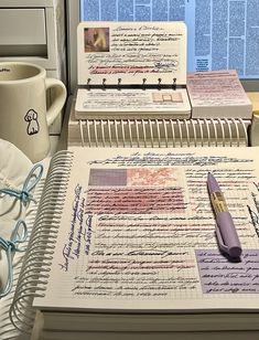 two notebooks with writing on them next to a cup and pen in front of a window