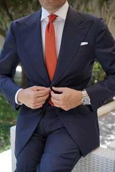 a man in a suit and orange tie sitting on a white chair with his hands under his lapel