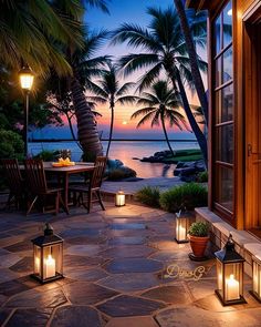 an outdoor patio with lanterns and palm trees at dusk, overlooking the water's edge