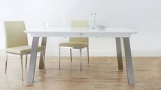 a white table with two beige chairs and a vase on the top, in front of a white wall