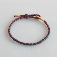 a red, green and gold string bracelet on a white surface with a golden bar