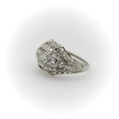 This platinum diamond ring is on the cusp of the Edwardian and Art Deco eras. The centerpiece of the ornate ring are three Peruzzi cut bezel set diamonds —one is an elongated rectangular shape, while the diamonds on either side are truncated, cut specifically for this ring. The larger diamonds are surrounded by a selection of 40 single and Old Mine Cut diamonds that are bead set into the ring. The ring has layers divided by milgrain work that tie together the lace-like patterns of the Edwardian era and the geometric designs of the Art Deco era in alternating bands. There is an added detail of a hand engraved scrolling foliate on each side of the ring. The ring has open work on the bottom that lets in the light and also reveals the triangular bottom shape of the diamonds—a nice touch that h Art Deco Emerald-cut Single Diamond Rings, Art Deco Asscher Cut Diamond Jewelry, Art Deco Three Stone Platinum Jewelry, Classic Jewelry With Baguette Diamonds In Rectangular Shape, Timeless Square Cut Rose Diamonds Jewelry, Heirloom Rectangular Brilliant Cut Rings, Rectangular Diamond White Ring With Baguette Diamonds, Formal Platinum Jewelry With Baguette Diamonds, Square Cut Diamond Ring With Rose Cut Diamonds