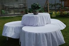 three round tables covered with white tablecloths in the grass near a building and potted plant