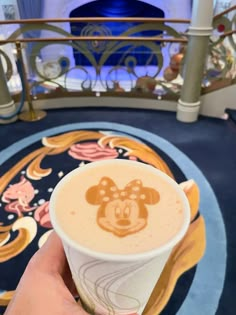 a hand holding up a foamy drink in front of a disney cruise ship staircase