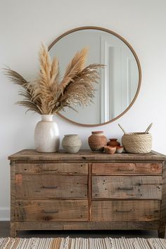 a wooden dresser topped with a mirror and vase filled with dried plants on top of it