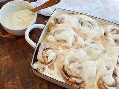 a pan filled with cinnamon rolls sitting on top of a wooden table next to a bowl of whipped cream