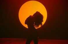 the silhouette of a woman standing in front of a large orange sun with her hair blowing back