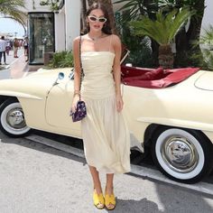 a woman standing in front of a car wearing sunglasses and a white dress with yellow heels