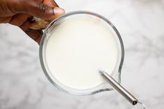 a person holding a glass with milk in it on a marble counter top next to a metal measuring spoon