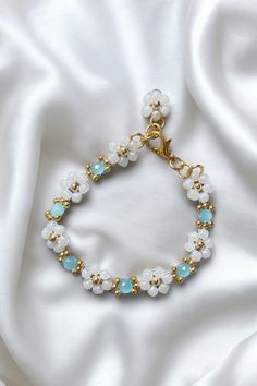 Coastal Breeze Daisy Chain Bracelet - 18K Gold-Plated, Aqua Blue Gem & White Glass Flower, Adjustable Chain, Gift for Her Add a touch of serene elegance to your jewellery collection with the Coastal Breeze Daisy Chain Bracelet, a beautiful piece that blends luxury with refreshing colour. Featuring delicate white glass flowers arranged in a daisy chain design, each flower is centred with shimmering 18K gold-plated beads and complemented by a vibrant aqua blue gem. This bracelet brings a sophistic Adjustable White Jewelry With Flower Decoration, Handmade White Crystal Bracelet With Flower Shape, Handmade White Crystal Bracelet In Flower Shape, Handmade White Crystal Flower Bracelet, Delicate White Beaded Round Bracelets, Adjustable White Bracelet With Flower Decoration, White Flower-shaped Wedding Bracelets, Light Blue Beaded Flower Jewelry, Delicate White Flower Bracelets