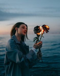 a woman holding a flower in front of the ocean at night with flames coming out of it