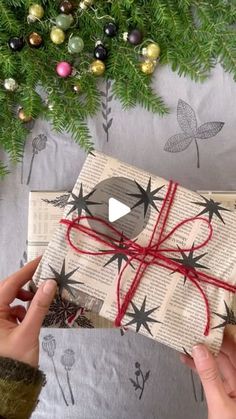 two hands holding a wrapped present in front of a christmas tree with ornaments on it