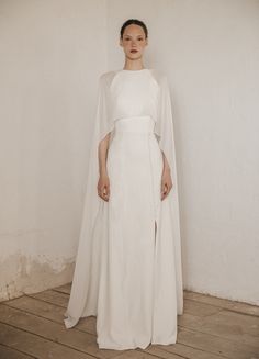 a woman standing in front of a wall wearing a white dress with cape over her shoulders