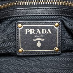 This Prada TessuTo handbag is made of black nylon canvas and has rolled leather indexes. The Bag is also decorated with gold -colored details. A golden Prada lettering shines on the front. The Bag Has a magnetic closure at the upper opening. Inside the Bag there are a zipper compartment and a slide compartment. The Bag can be in the hand or with a removable leather shoulder strap over the shoulder. Leather Bags With Gold-tone Logo Plaque And Double Handle, Leather Bags With Double Handle And Gold-tone Logo, Black Leather Bags With Logo Plaque, Gold Bag With Gold-tone Logo Plaque And Double Handle, Black Shoulder Bag With Gold-tone Logo For Everyday Use, Business Shoulder Bag With Gold-tone Logo Plaque, Luxury Double Handle Shoulder Bag With Gold-tone Logo Plaque, Gold Shoulder Bag With Logo Plaque For Everyday, Luxury Shoulder Bag With Logo Plaque And Top Handle