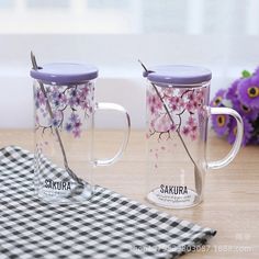 two glass mugs with flowers painted on them sitting on a table next to a gingham cloth