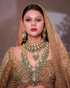 a woman wearing a gold and green bridal outfit with jewelry on her neck, in front of a wall