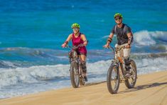 two people riding bikes on the beach near the ocean