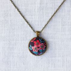 Inspired by Liberty Art Fabric, I developed my collection of vintage-style jewellery. The vibrant floral motifs of Liberty fabric are truly celebrated and showcased in this collection. A great gift for nature-lovers that will never go out of fashion. This necklace features red flowers in a dark background. It has an antique bronze alloy pendant tray and an oval pendant. The long brass chain has a lobster clasp and a tiny bee beside it. Pendant 2.8 x 2.8 cm Handmade in my studio based in England. Vintage Flower Charm Necklace For Gift, Flower Shaped Necklace With Vintage Charm For Gift, Flower Shaped Necklace With Vintage Charm As Gift, Handmade Vintage Jewelry With Round Pendant, Vintage Necklaces With Flower Charm Round Pendant, Vintage Flower Charm Pendant Jewelry, Floral Print Round Jewelry Gift, Vintage Necklace With Flower Charm Round Pendant, Vintage Pendant Necklace With Flower Charm