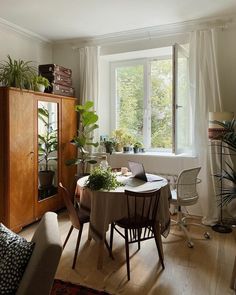 a living room filled with furniture and a table next to a large window covered in potted plants