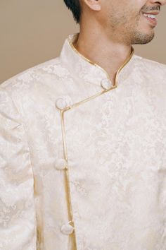 A Vietnamese ao gam jacket fit for a modern groom. This men’s Vietnamese wedding outfit features an elegant and refined design made with ivory and light gold dragon brocade fabric. Pair this with one of our beautiful ao dais for the perfect modern Vietnamese-American wedding tea ceremony. Bride's Dress Recommendation: Pairs well with our white and gold Kathy Vietnamese Wedding Dress. This jacket is named after the Tran Dynasty that ruled between 1225-1400, during which time they resisted invader Elegant Traditional Wear With Stand Collar For Wedding, Elegant Nehru Jacket For Traditional Ceremonies, Gold Long Sleeve Sherwani For Groom, Elegant Gold Sherwani For Traditional Ceremonies, White Stand Collar Bandhgala For Wedding, White Bandhgala With Stand Collar For Wedding, Elegant Gold Nehru Jacket For Groom, Gold Long Sleeve Bandhgala For Groom, Gold Brocade Outerwear For Wedding