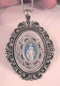 This silvertone necklace features a lovely small-to-mid-sized pendant with a swoop scalloped outer rim that gives it charm and character.  The center is accented with a blue finished floral medal inset with the image of The Immaculate Conception, Our Lady of Grace -  The Miraculous Mary.  It is inscribed with the Miraculous Prayer and the pendant has a shiny finish on the reverse.  It measures 1 5/8 inches wide and 2 3/8 inches long.  The chain is 24 inches and closes with a safety catch. Silver Necklaces With Oval Pendant Vintage Charm, Silver Necklace With Oval Pendant And Vintage Charm, Antique Silver Medallion With Miraculous Medal, Vintage Silver Necklace With Miraculous Medal, Vintage Miraculous Medal Jewelry, Our Lady Of Grace, Cross Medal, Lady Of Lourdes, Our Lady Of Lourdes