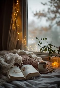 an open book and cup on a bed next to a window with lights in the background