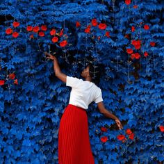 a woman standing in front of a blue wall with red flowers on it and reaching up