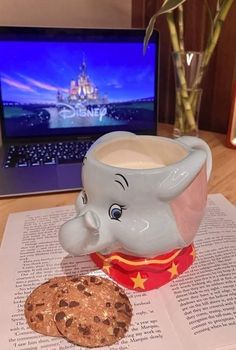 an elephant mug sitting next to a cookie on top of a book with a laptop in the background