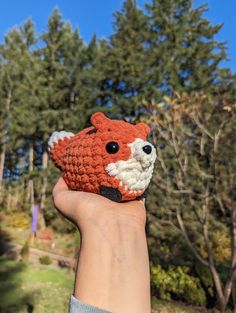 a hand holding up a small crocheted fox head in front of some trees