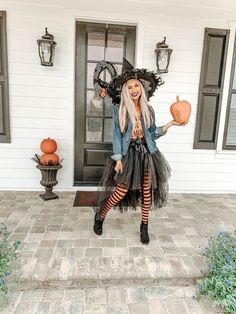 a woman dressed as a witch standing in front of a house holding two pumpkins