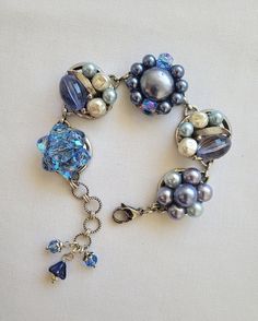 This bracelet has been made from 5 vintage clip earrings! The earrings I've chosen are in a medium blue and purple (with a bluish tone); all set in silver. The pieces are securely attached to a silver tone bracelet base. It's adjustable from 7 to 8 inches long and has 2 blue crystals and a purple glass flower charm that dangle down the chain. A great gift for a bridesmaid or for Mother's Day  The bracelet will arrive wrapped in tissue and in a silver gift bag.  The package will be mailed USPS,  Ground Advantage with tracking and insurance,  in a small cardboard box. Blue Metal Clip-on Jewelry, Metal Clip-on Jewelry For Jewelry Making, Blue Vintage Nickel Free Bracelets, Blue Beaded Metal Jewelry, Vintage Blue Nickel-free Bracelets, Blue Metal Clip-on Earrings, Blue Bracelet With Unique Variations, Vintage Blue Nickel-free Bracelet, Blue Round Clip-on Jewelry