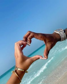 two hands making a heart shape with their fingers on the beach, one holding a cell phone