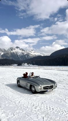 two people are sitting in an open convertible car on snow covered ground with mountains in the background