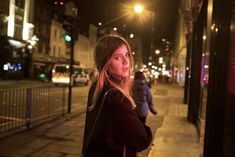 a woman standing on the sidewalk at night with her hand in her pocket and looking off into the distance