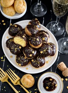 a plate full of chocolate covered donuts next to some champagne glasses and gold forks