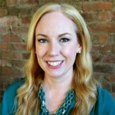 a woman with long blonde hair wearing a green shirt and smiling at the camera while standing in front of a brick wall