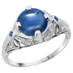 Indulge in timeless elegance with this exquisite vintage cocktail ring, a true masterpiece of fine jewelry craftsmanship. Featuring a stunning GIA Certified No Heat Burma Cabochon Round Sapphire, weighing an impressive 2.23 carats, this ring is a radiant symbol of sophistication and luxury. The captivating sapphire is set in a beautifully engraved and millgrain platinum band, exuding a vintage charm that is both unique and enduring. Main Features: Center Stone: Type: GIA Certified No Heat Sapphire Shape: Cabochon Round Weight: 2.23 Carats Color: Deep, Rich Blue Accent Stones: Diamonds: Total Weight: 0.15 Carats Cut: Round Brilliant Setting: Pave, enhancing the ring’s sparkle Side Sapphires: Shape: Square Color: Matching deep blue, perfectly complementing the center stone Ring Setting: Meta Yellow Sapphire Rings, Vintage Cocktail Ring, Round Sapphire, Platinum Diamond Rings, Gold Cocktail Ring, Diamond Cocktail Rings, Platinum Ring, Blue Accents, Exquisite Jewelry