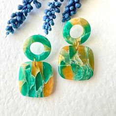 two green and yellow earrings sitting on top of a white table next to blue flowers