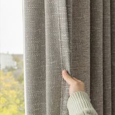 a person's hand is holding the curtain in front of a window with grey curtains