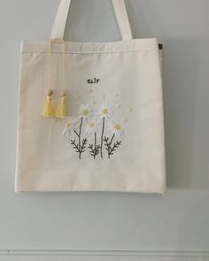 a white tote bag with daisies and tassels hanging on the wall