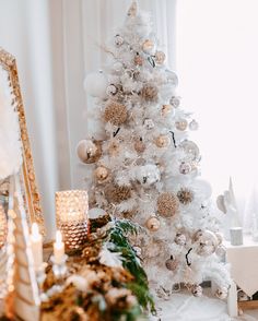 a white christmas tree with gold and silver ornaments in front of a mirror, candles and other holiday decorations