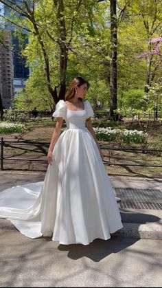 a woman in a white dress is standing on the sidewalk with trees and buildings behind her