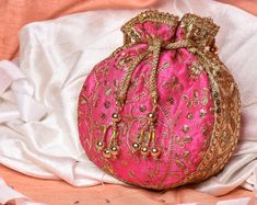 Pink Potli Bag with Pearl Handle Strap With its classy zari work and a pearl handle strap all in gold, this Pink Potli Bag is ideal for this wedding season. Fabric: Raw Silk Color: Pink Embroidery: Zari Work in Gold Size & Dimensions: Height - 8 inWidth - 7.5 inDepth - 1.5 in Strap: Pearl Handle Strap Closure: Tasseled Drawstring Compartments: Single compartment Care: Wipe with a soft, clean & dry cloth to remove dust When not in use, store it in a dust bag for longer product life Avoid Traditional Cheap Pink Bag, Bridal Mask, Potli Bag, Ethnic Bag, Wedding Bags, Pink Embroidery, Potli Bags, Embroidery Bags, Royal Blue And Gold