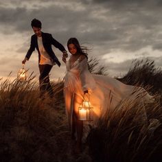 a man and woman walking through tall grass with lanterns in their hands as the sun goes down