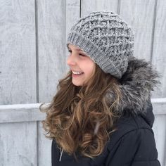 a woman wearing a gray knitted hat with a fur pom in front of a wooden fence