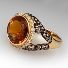 The ring is centered with one (1) round mixed cut natural citrine set into a bezel. The center stone is bordered with twenty-four (24) bead set, round brilliant cut diamonds. The ring is also accented with one hundred (100) round brilliant cut champagne colored diamonds bead set into black rhodium topped settings. The ring measures 15.4mm at the top, rises 8.0mm above the finger, tapering to 3.0mm wide and 1.2mm thick at the base of the shank. It is currently a size 7.25. Elegant Amber Rings With Gemstone Accents, Luxury Round Gemstones With Diamond Accents, Luxury Citrine Topaz Ring With Gemstone Accents, Yellow Gold Topaz Ring With Gemstone Accents, Luxury Round Cut Gemstones With Diamond Accents, Elegant Citrine Gemstones With Gemstone Accents, Luxury Citrine Gemstones With Accents, Luxury Brown Formal Rings, Luxury Citrine Jewelry With Diamond Accents