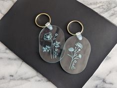 two glass key chains sitting on top of a black surface with flowers etched on them