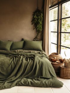 a bed with green sheets and pillows in front of a window next to a basket