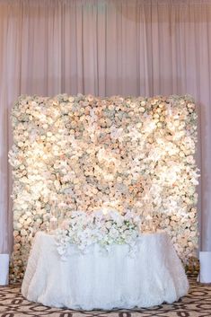 a white table with flowers on it in front of a wall that has lights and curtains